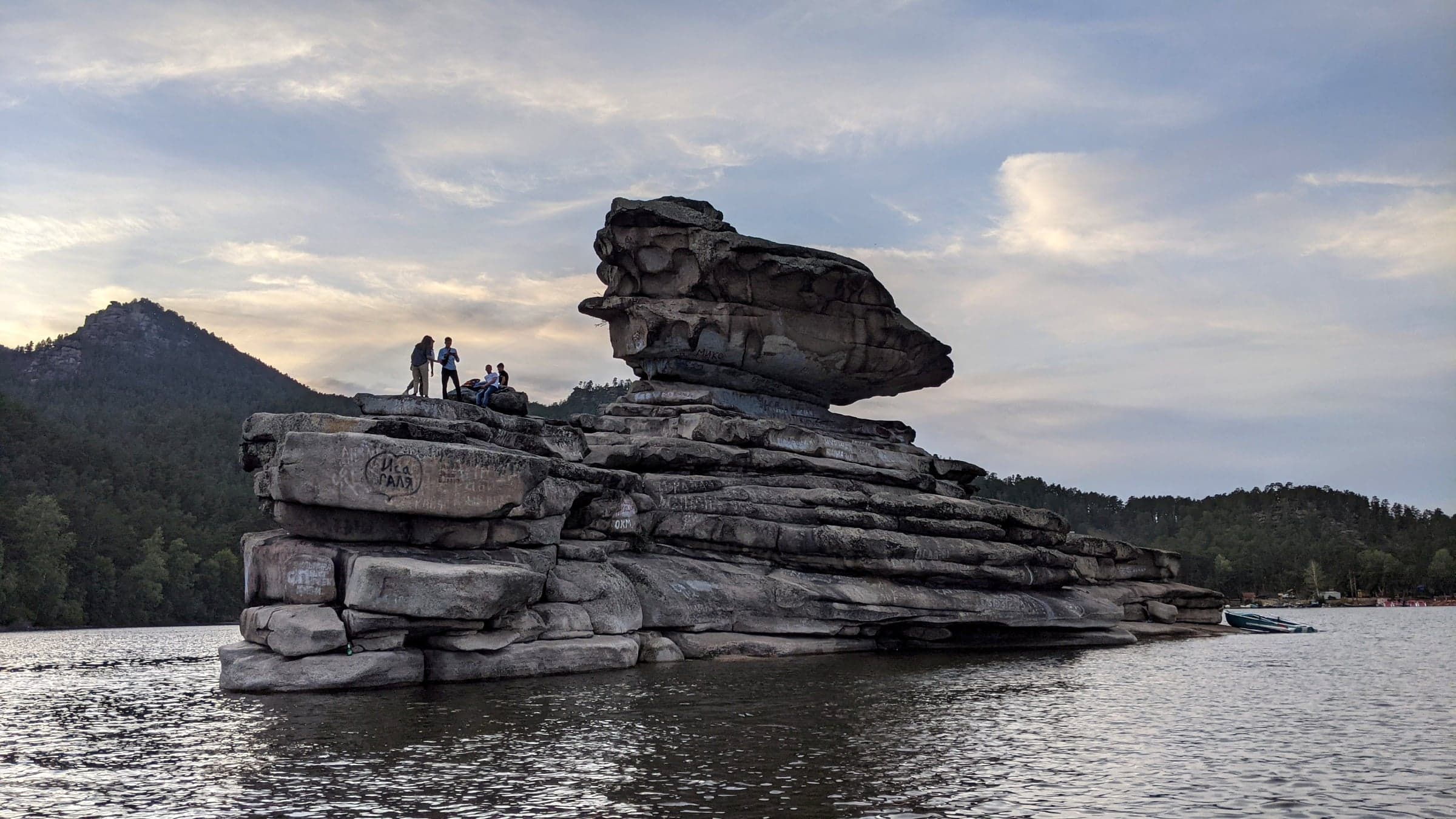 The Zhumbaktas balancing rock formation rising out of Burabay Lake at dusk, with silhouetted visitors standing on top and mountains in the background