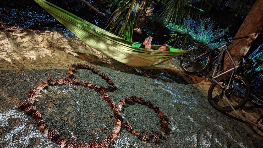 A hammock strung between pines lit by string lights at night, with a heart shape made of pine cones on the ground beside two mountain bikes