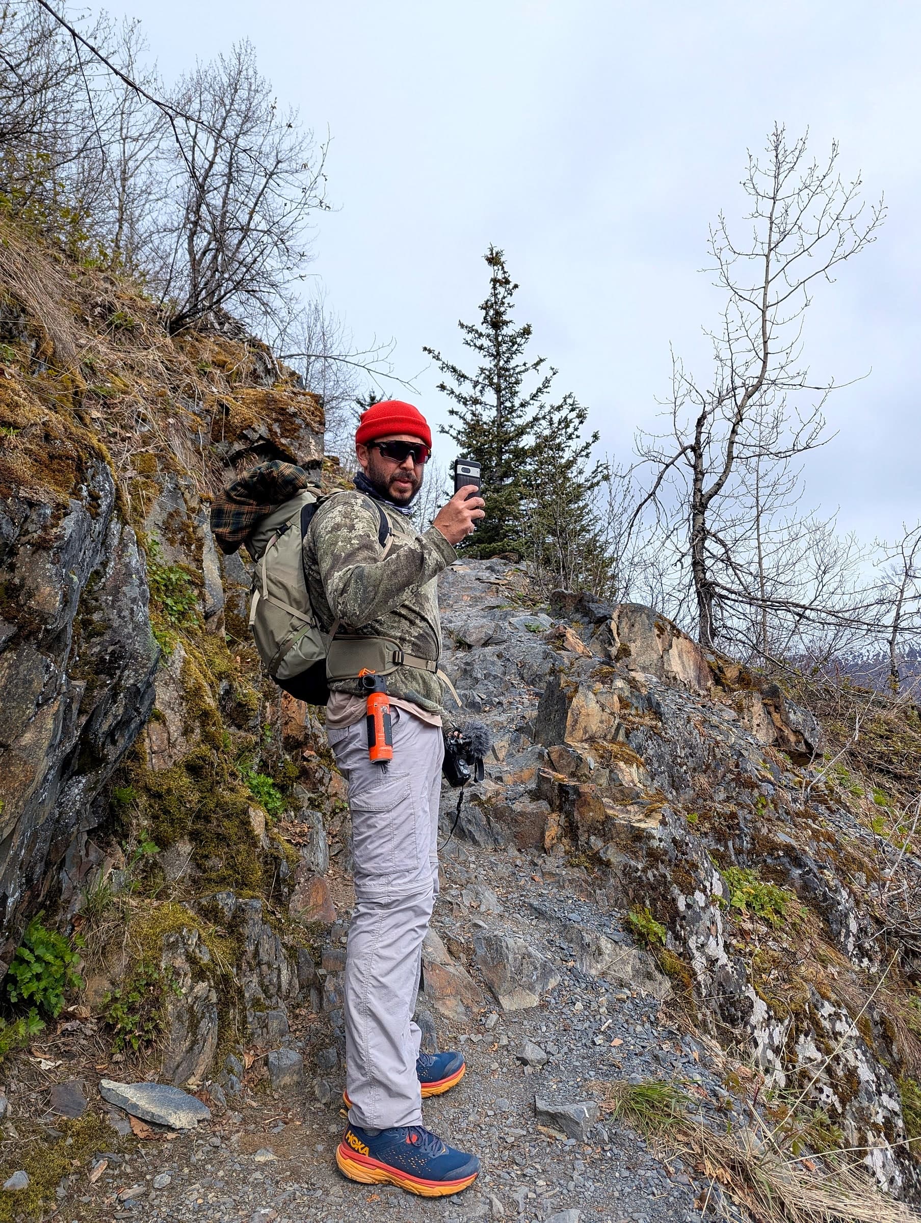 A hiker with a canister of bear spray clipped to his belt, standing at an overlook of Exit Glacier on the Harding Icefield Trail in Alaska
