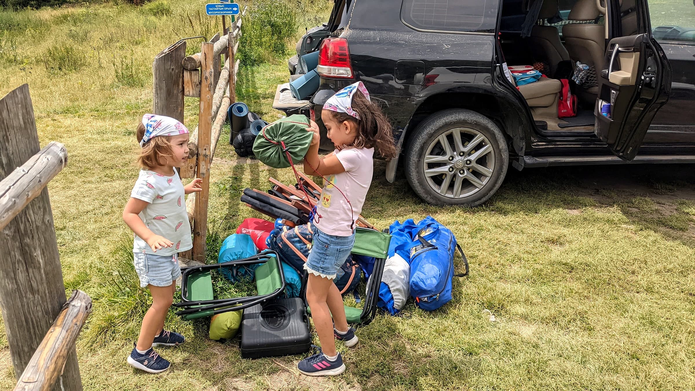 Two young girls loading camping chairs and sleeping bags into the back of a car at a campsite