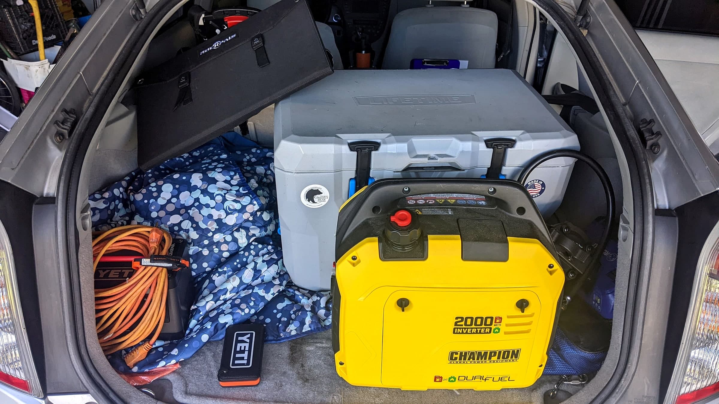 A yellow Champion 2000-watt dual-fuel inverter generator packed in a car trunk next to a gray Lifetime cooler