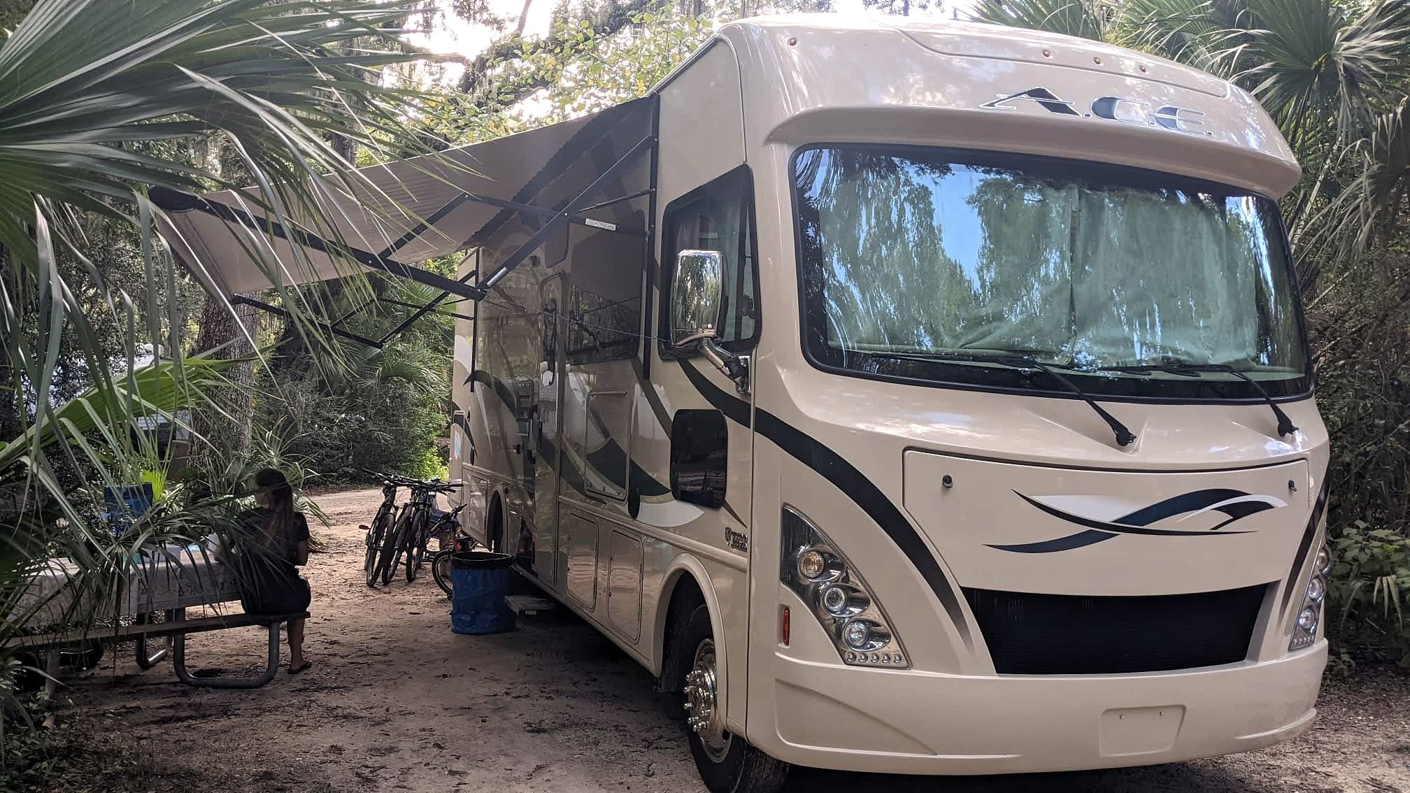 A rented Cruise America Class C RV parked at a wooded Florida campsite with an awning extended and bikes leaning against it