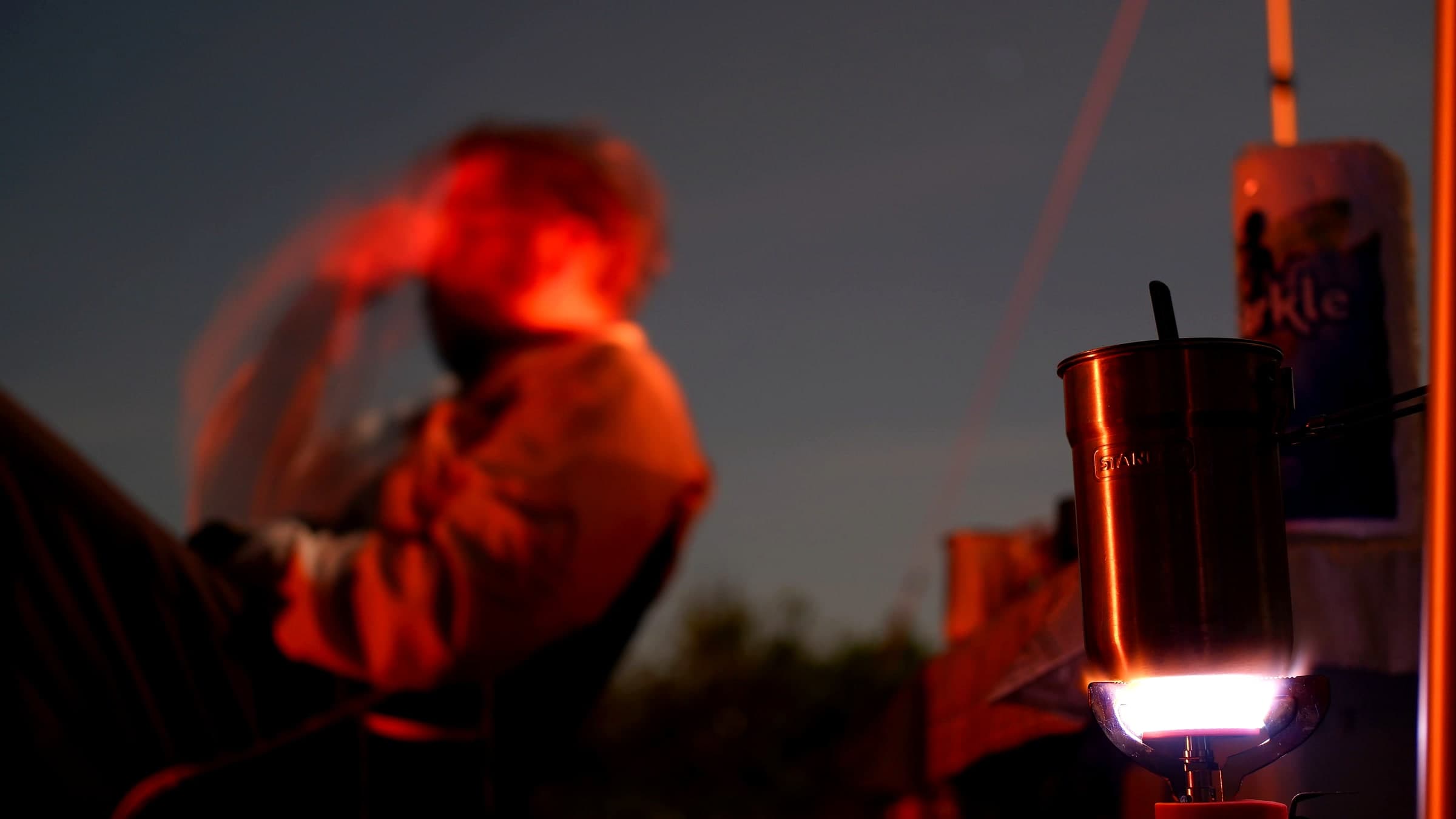 A green Coleman propane camp stove burning at night with a Stanley mug set on the grate