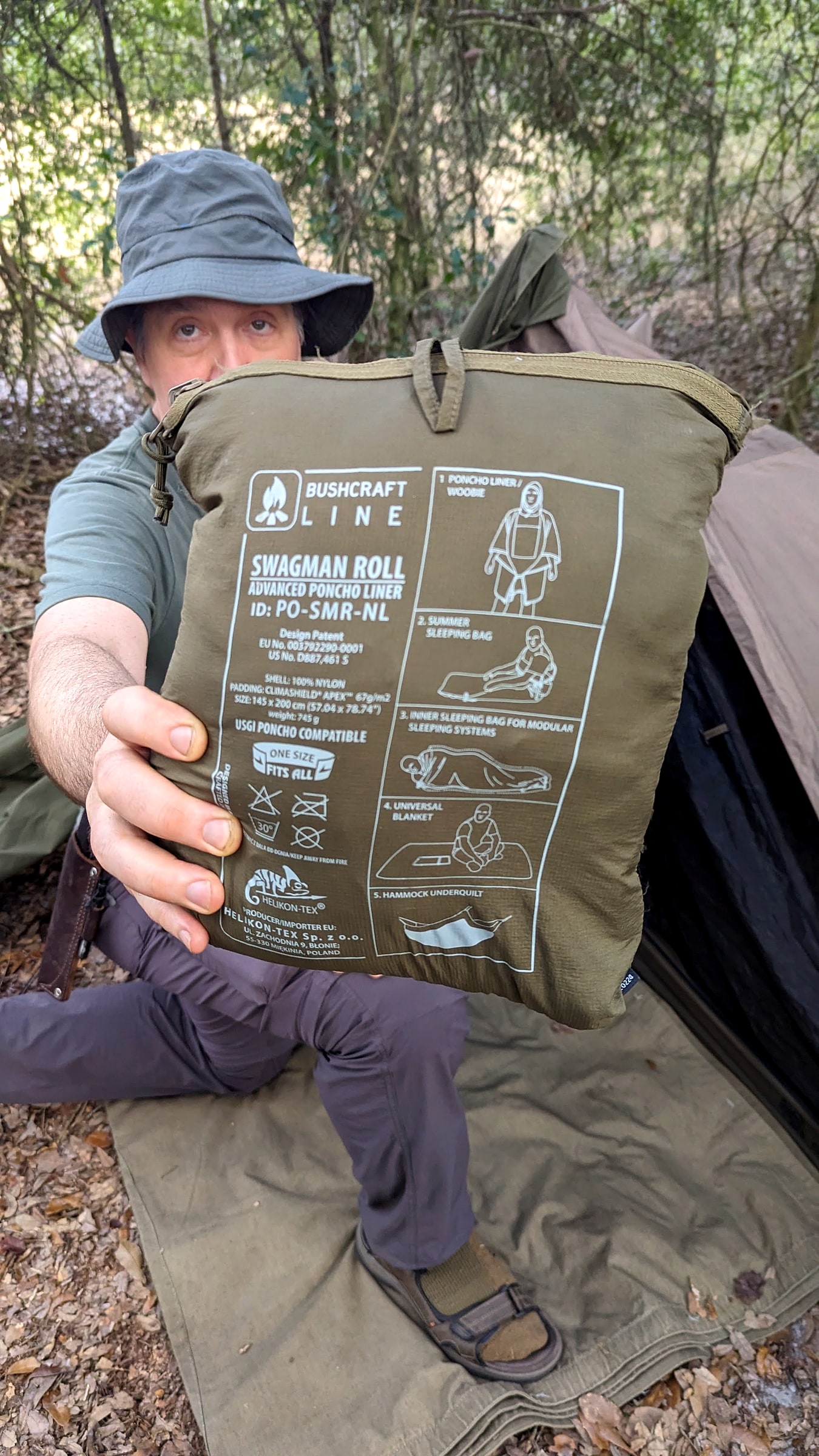 A man holding a rolled Helikon-Tex Swagman Roll poncho liner in a wooded backcountry campsite