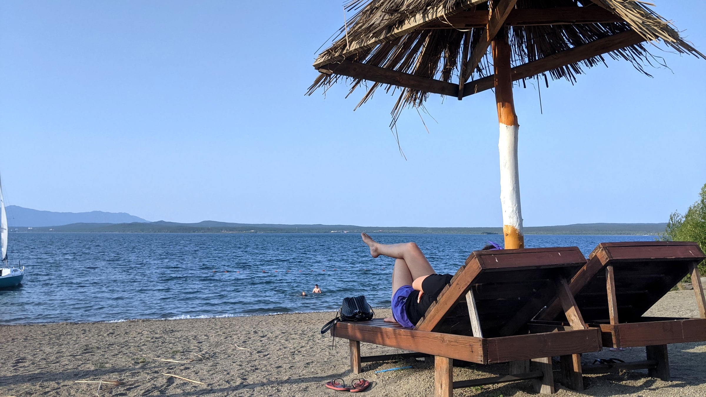 A wooden lounge chair under a thatched-roof beach umbrella on the sandy shore of Burabay Lake, with sailboats and swimmers visible on the water