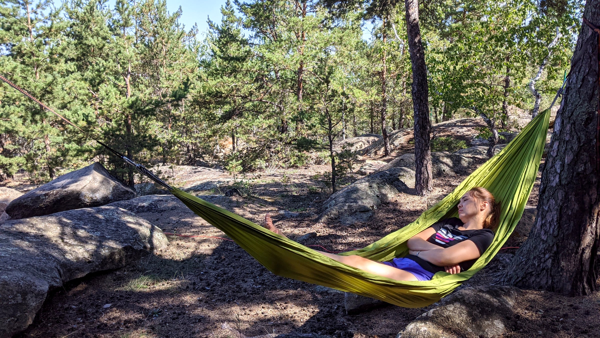 A green hammock strung between pine trees on a granite outcrop, with someone resting inside it in dappled forest light