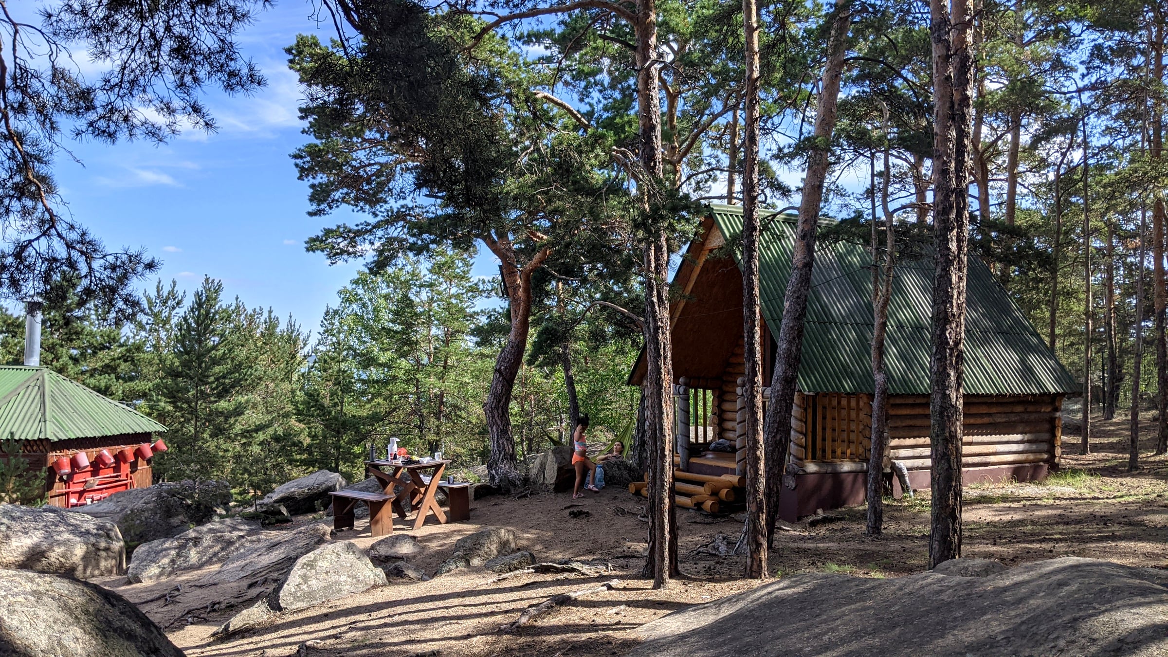 A-frame log cabins with green metal roofs tucked among tall pines at the Burabay glamping site, with a picnic table and a hammock strung nearby