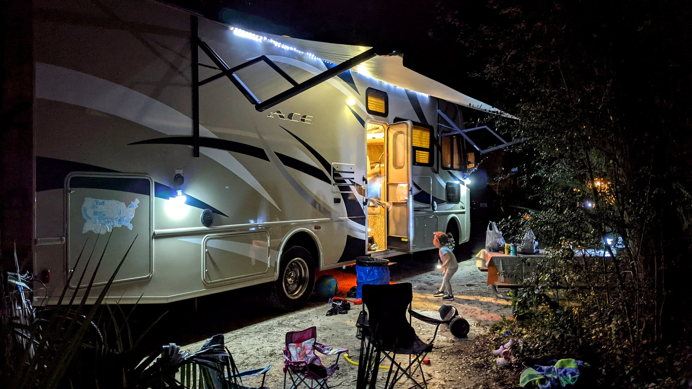 A rented Cruise America Class C RV parked and lit up at a campsite at night, with a child playing outside