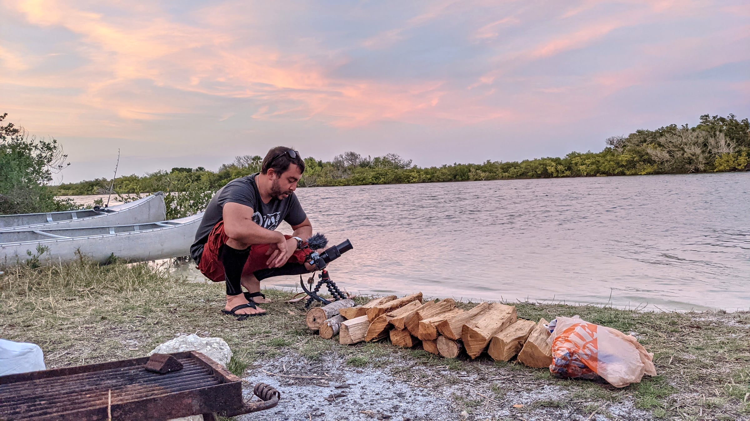 A man crouched by the shoreline at sunset next to a stacked pile of firewood, a tripod-mounted camera, and a beached aluminum canoe at Canaveral National Seashore
