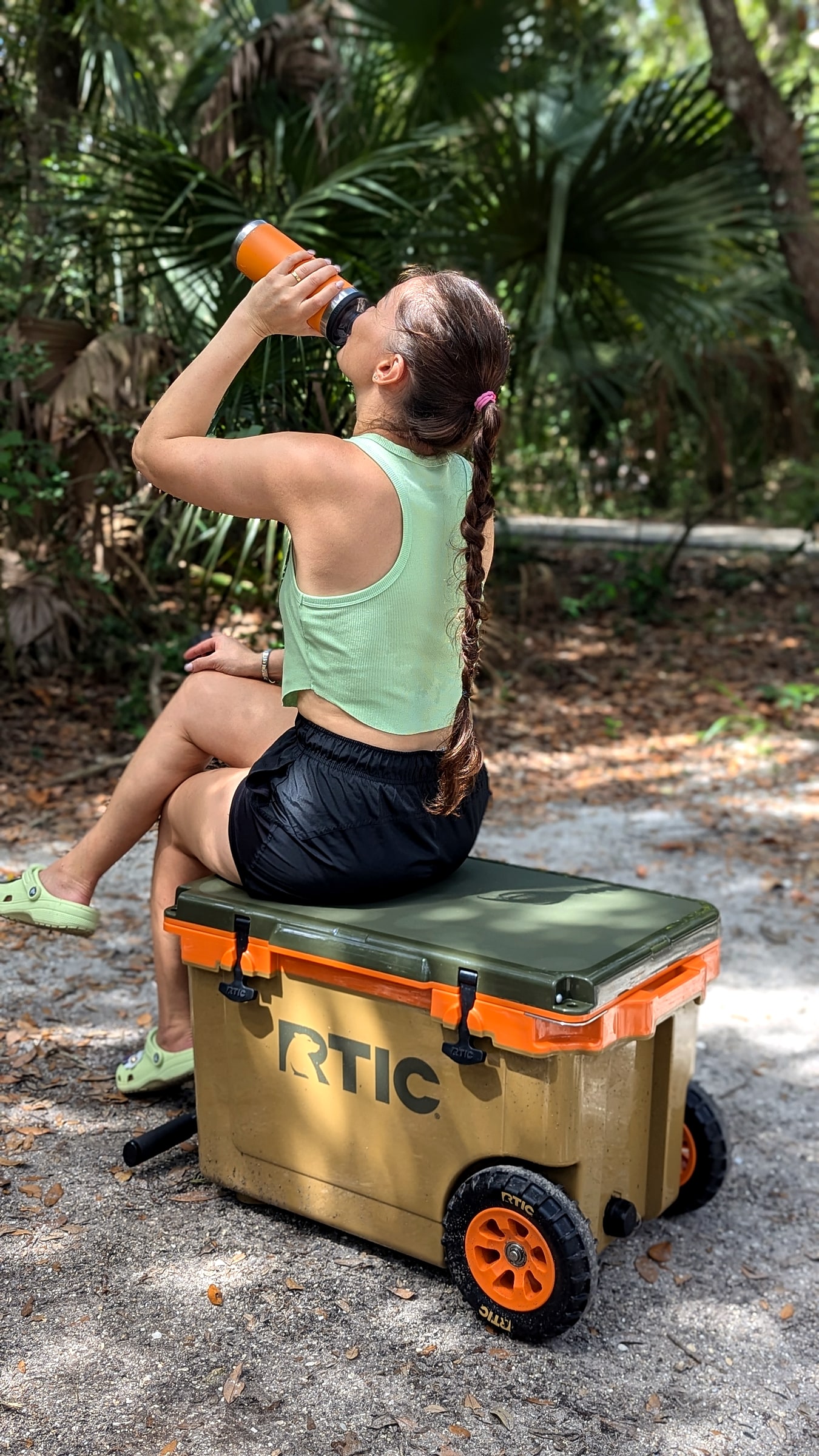 A woman sitting on a green and orange RTIC wheeled cooler at a campsite, drinking from an insulated tumbler