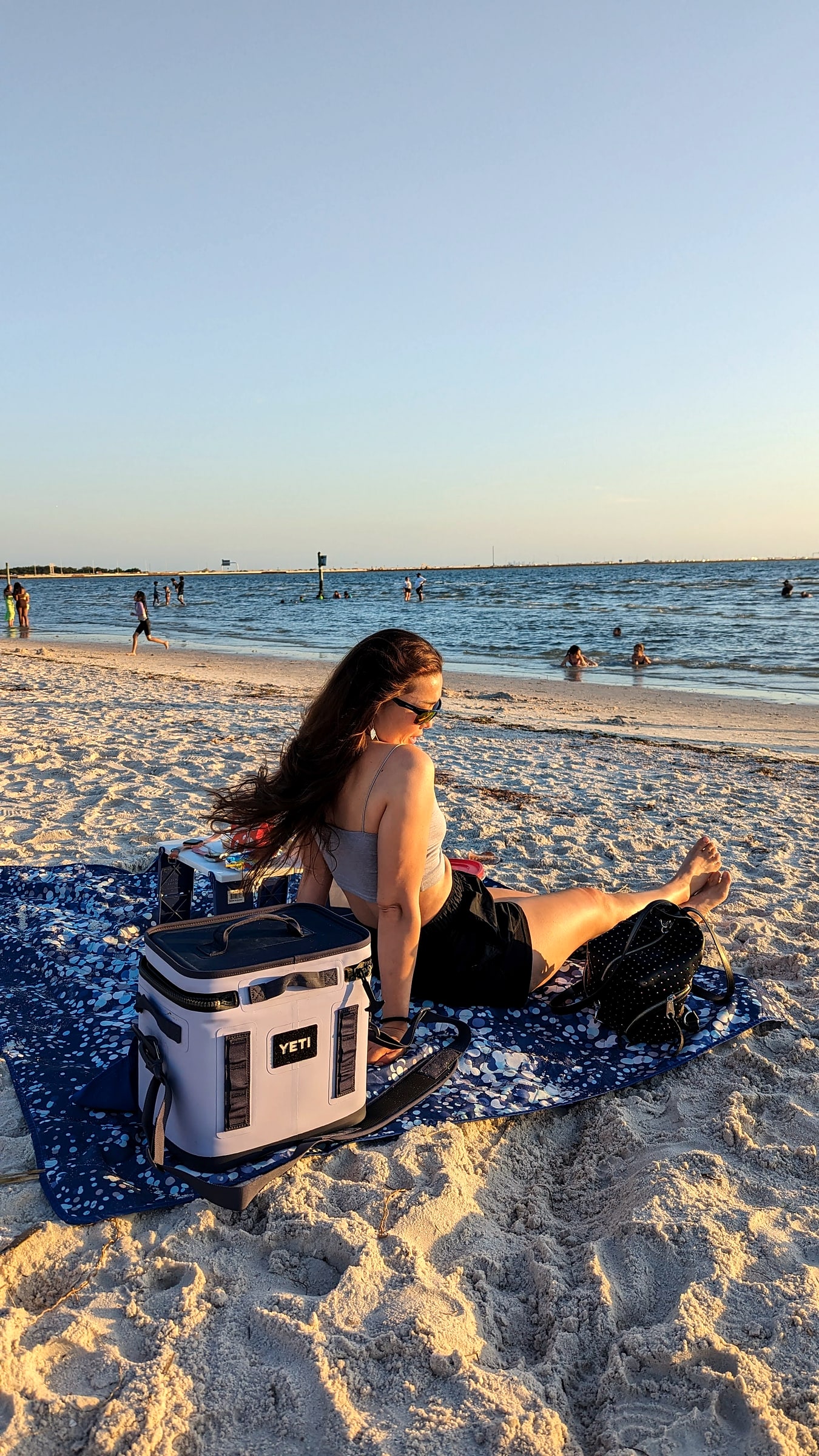 A Yeti Hopper soft cooler on a beach towel at sunset with people swimming in the background