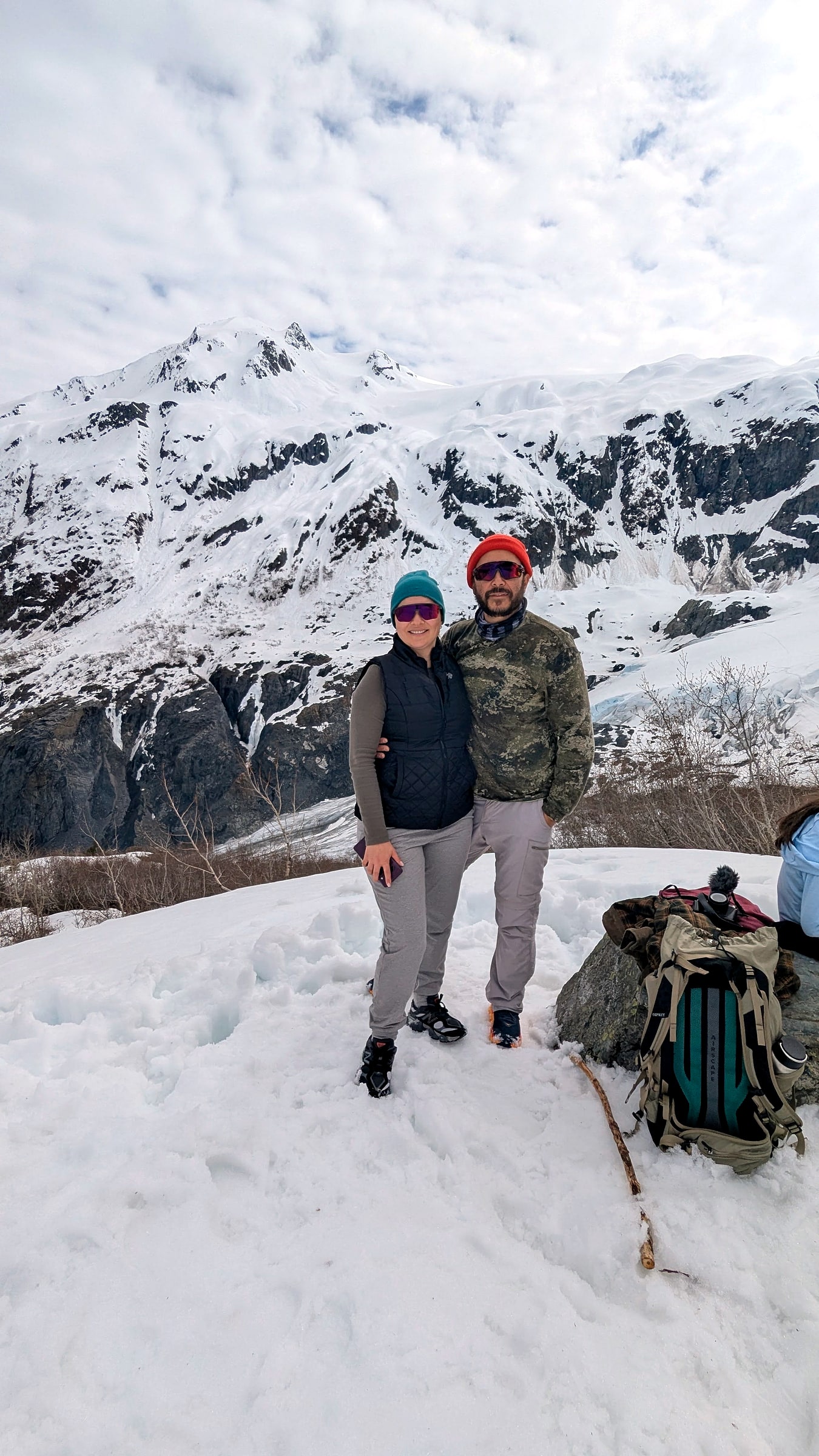 Two hikers standing at a snowy overlook on the Harding Icefield Trail with mountains in the background
