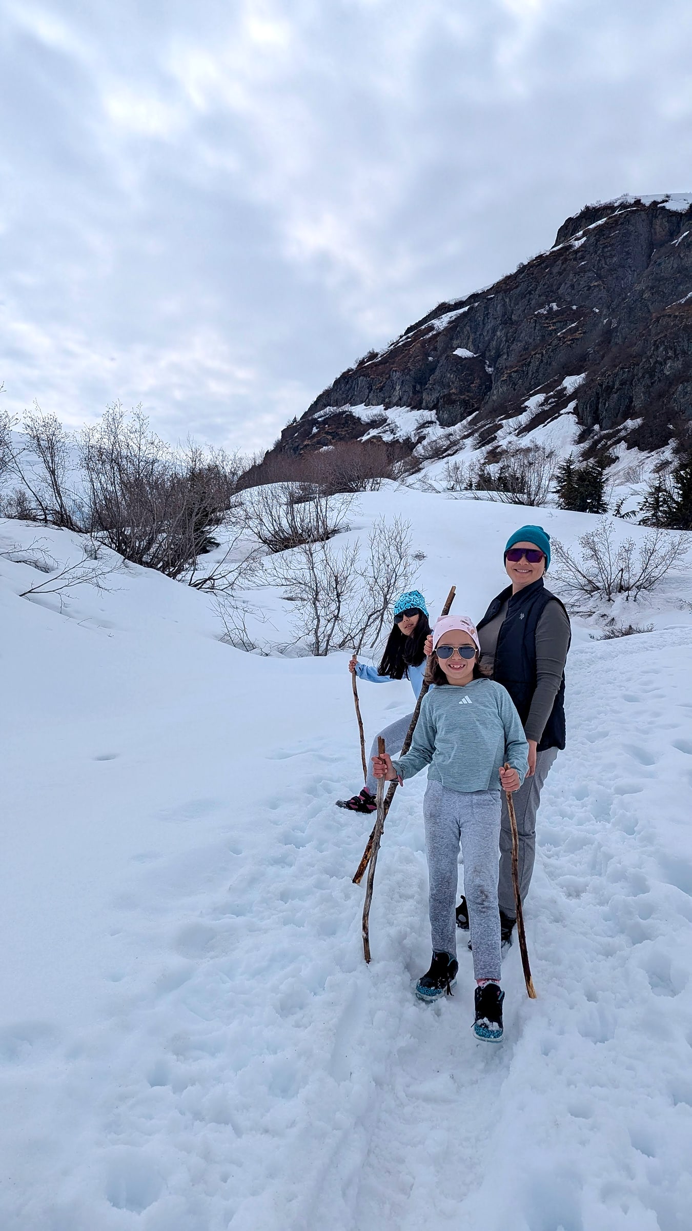 A parent and child hiking through a snowy patch on the Harding Icefield Trail, using a found stick as a walking pole