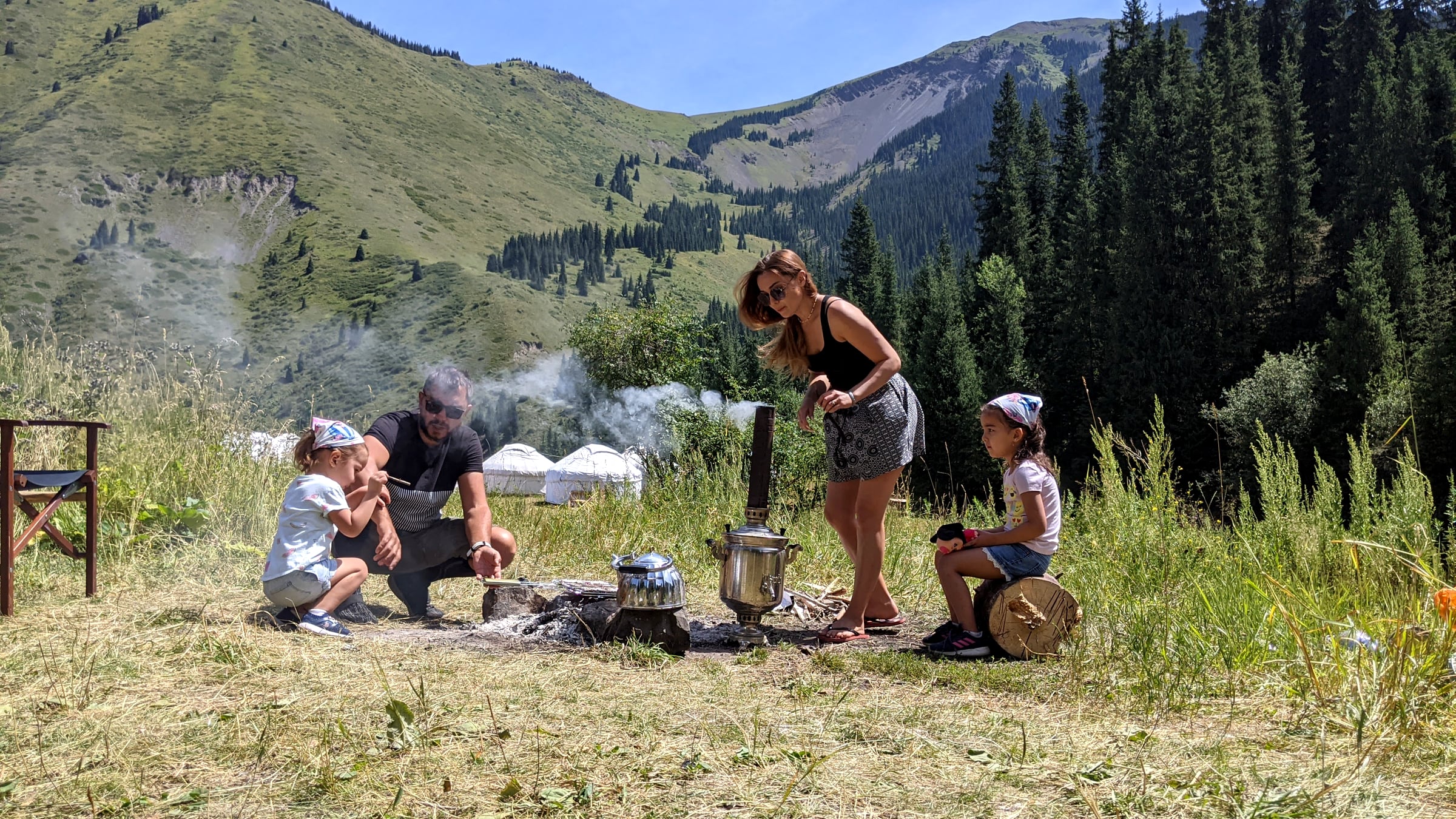 A samovar and kettle steaming over a campfire with mountains in the background, with a family gathered around brewing tea at a Kolsay Lakes campsite