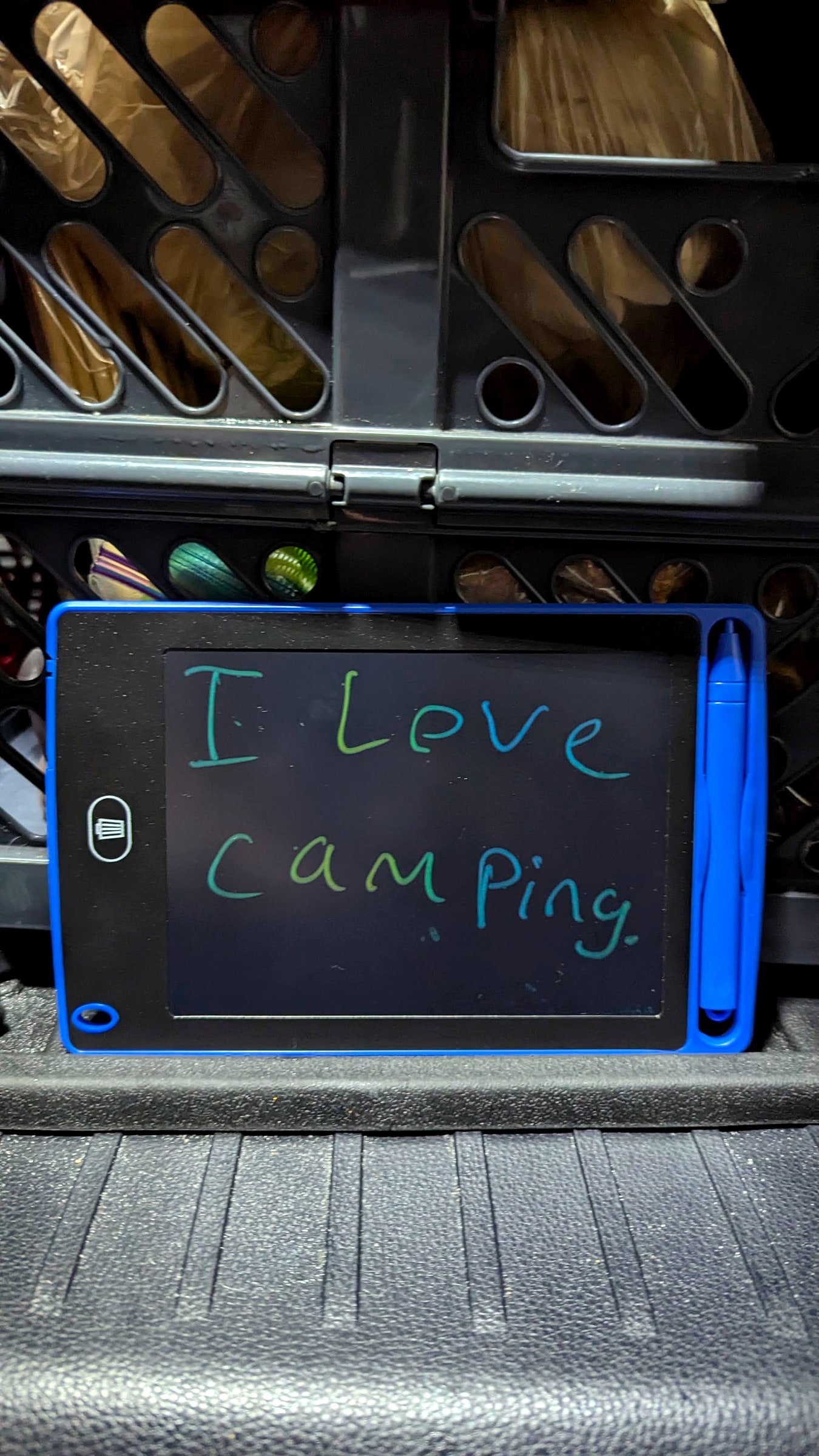 A child's LCD writing tablet reading 'I love camping' propped in the back of a packed car