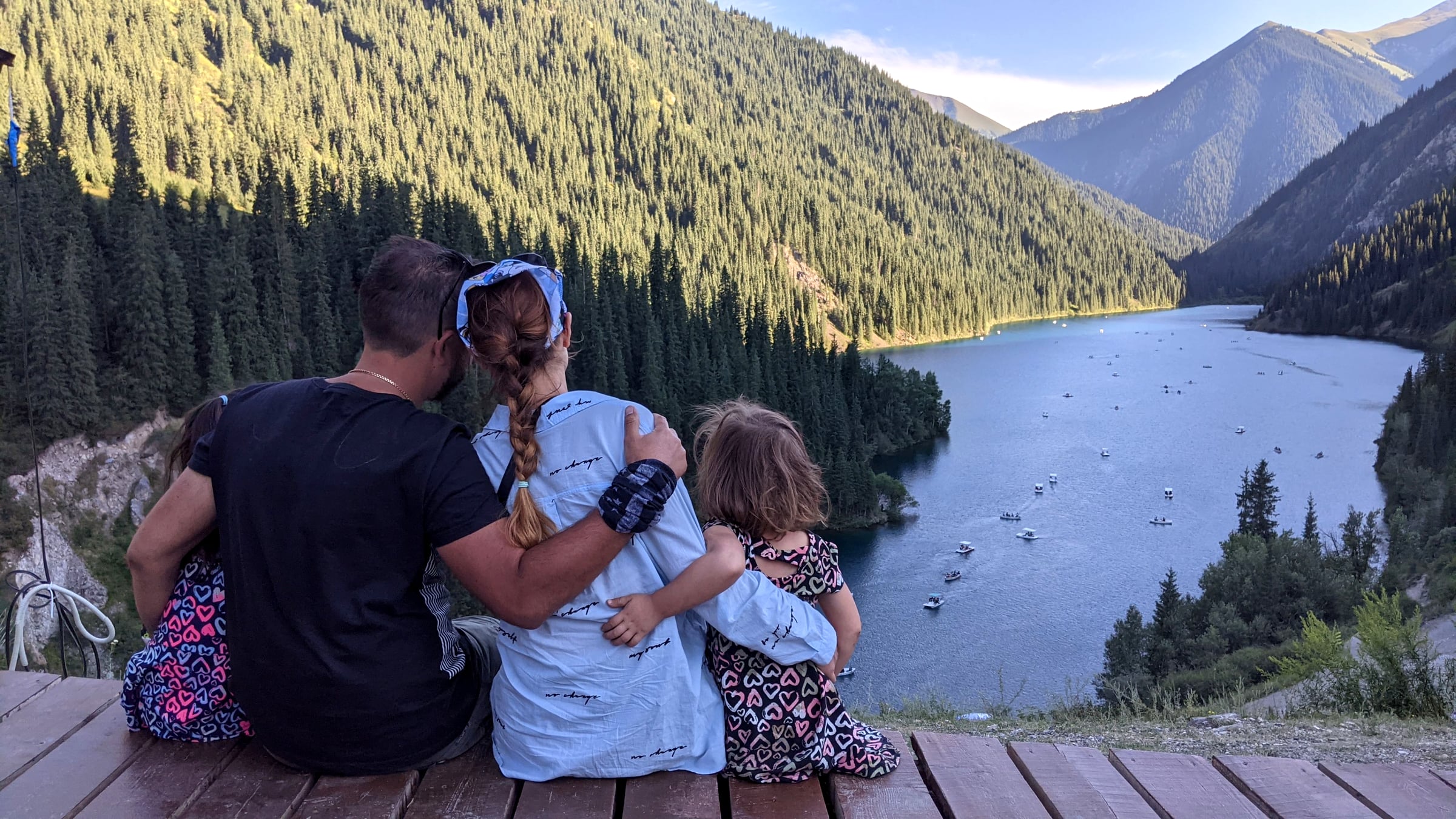 A family sitting on a wooden overlook deck above Kolsay Lake, watching small boats cross the deep-blue water surrounded by pine-covered mountains