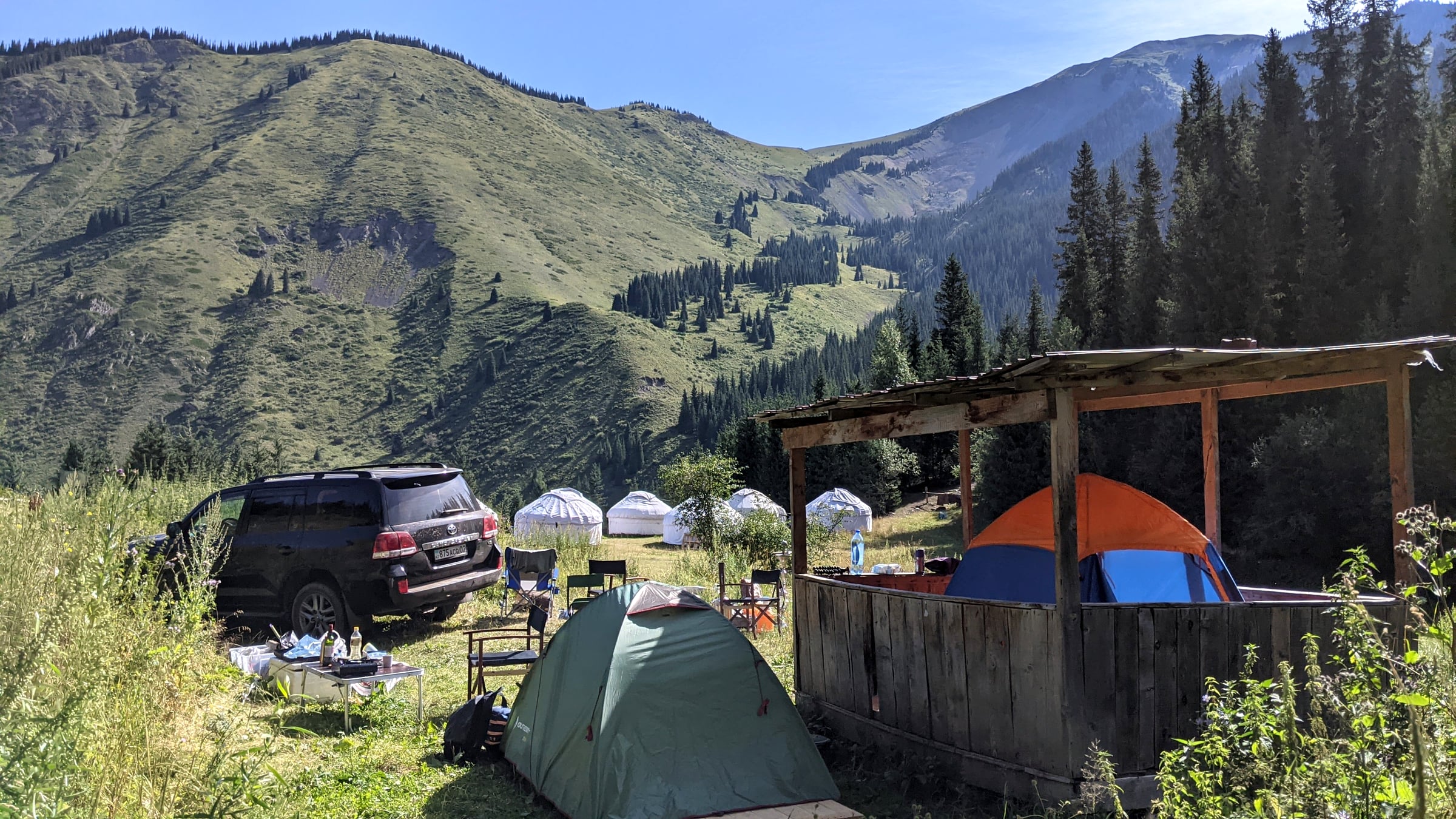 A mountain valley campsite with dome tents, a parked SUV, and a row of traditional white yurts set against steep green slopes near Kolsay Lake