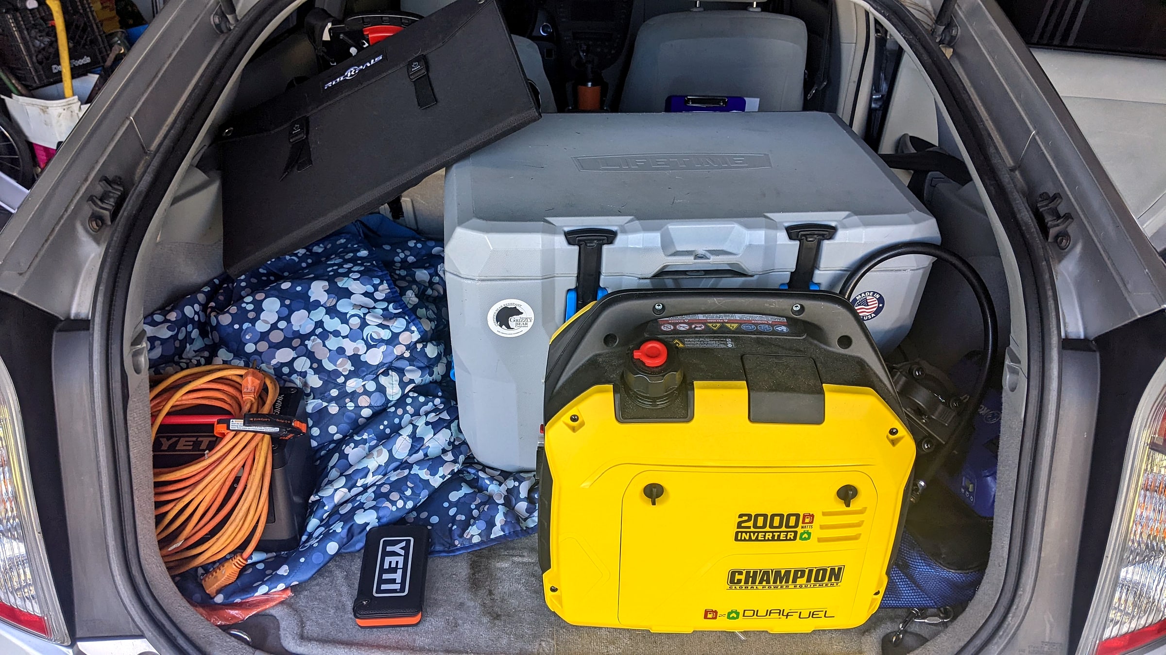 A yellow Champion 2000-watt dual-fuel inverter generator packed in a car trunk with camping gear