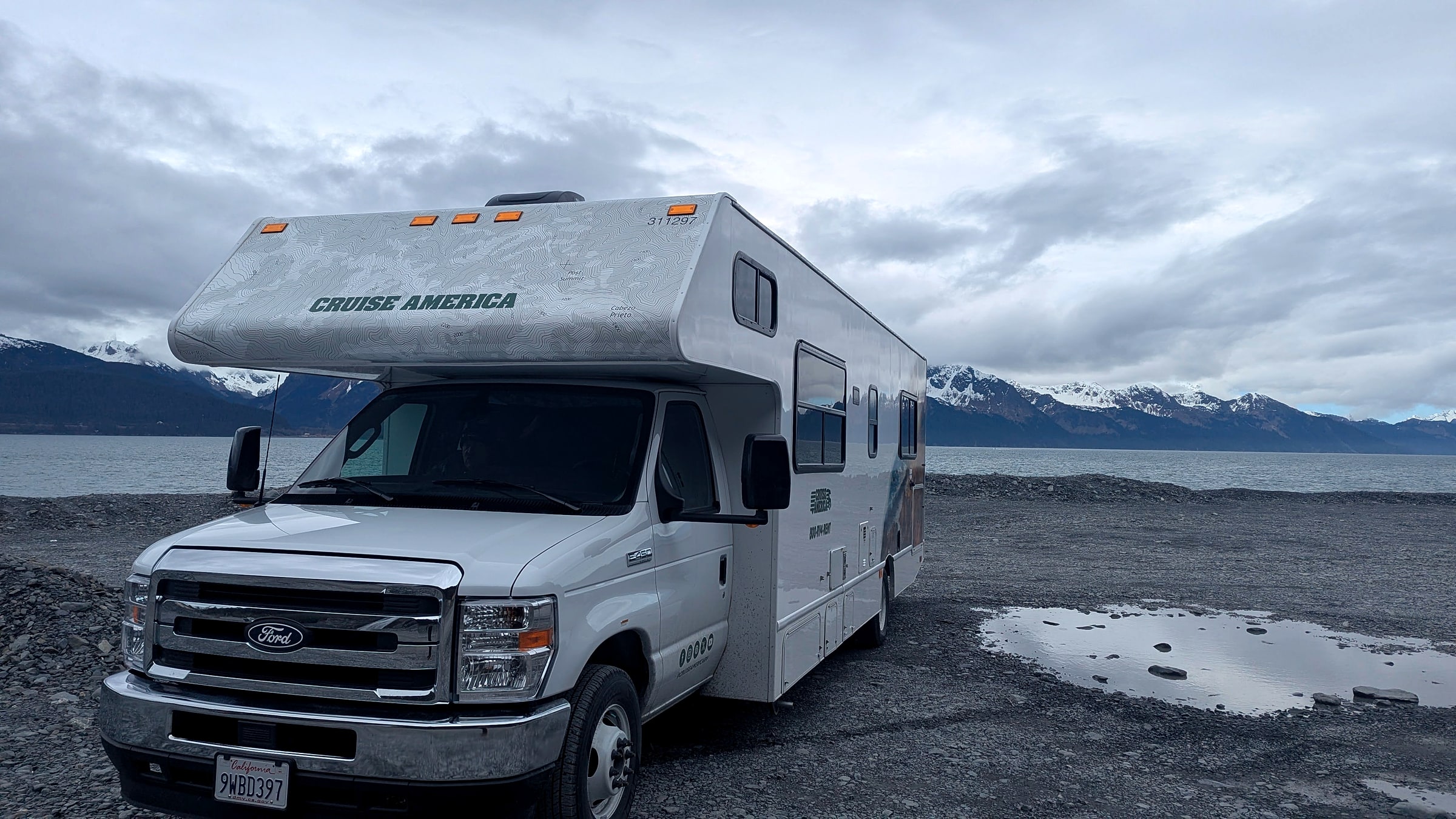 A rented Cruise America Class C RV parked on a gravel pullout along the coast in Alaska, with snow-capped mountains in the background