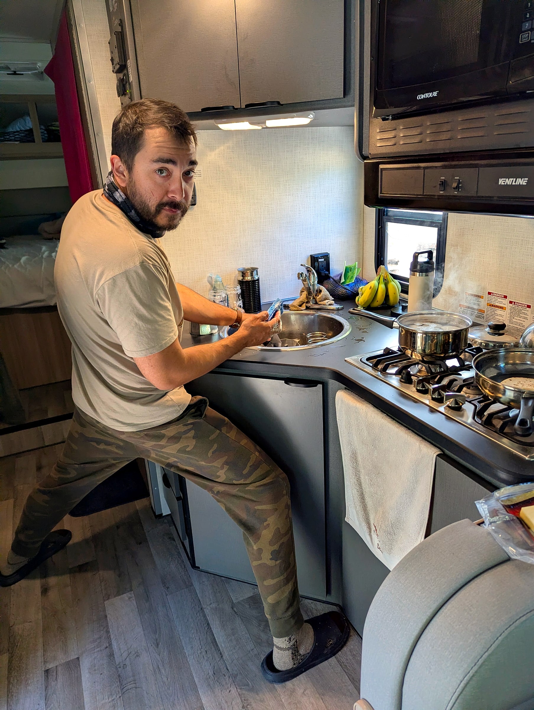 A man washing dishes at the small kitchen sink inside a rented RV in Alaska, with a pot of water boiling on the stove