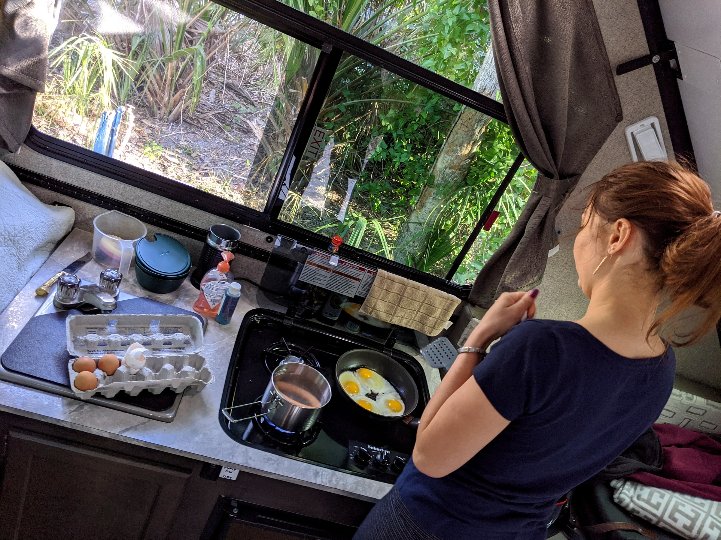 A woman cooking eggs on a small stovetop inside an RV galley kitchen, with an egg carton and coffee supplies on the counter