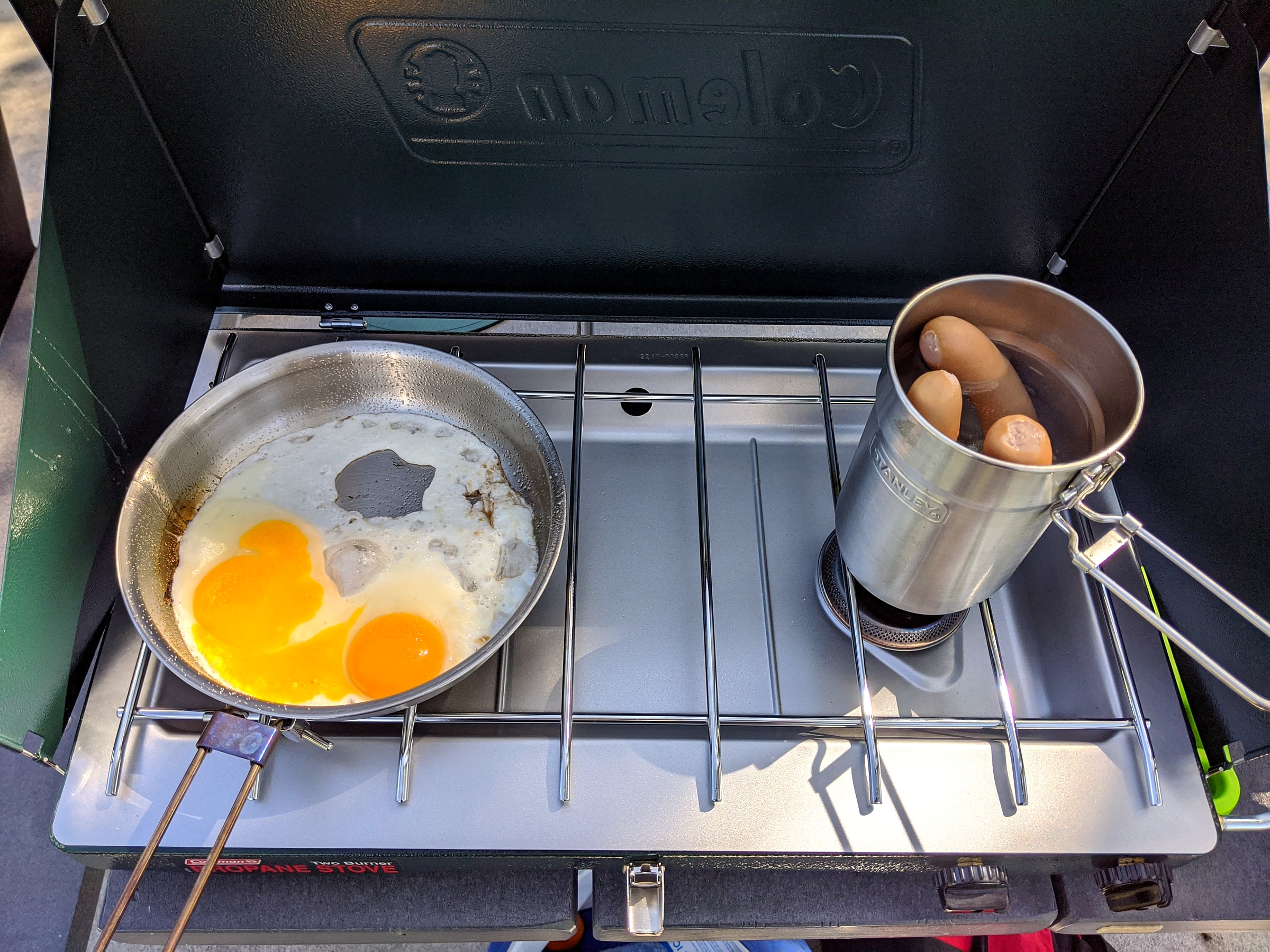 A green Coleman propane camp stove cooking eggs and hot dogs at a picnic table, with a spring water bottle beside it