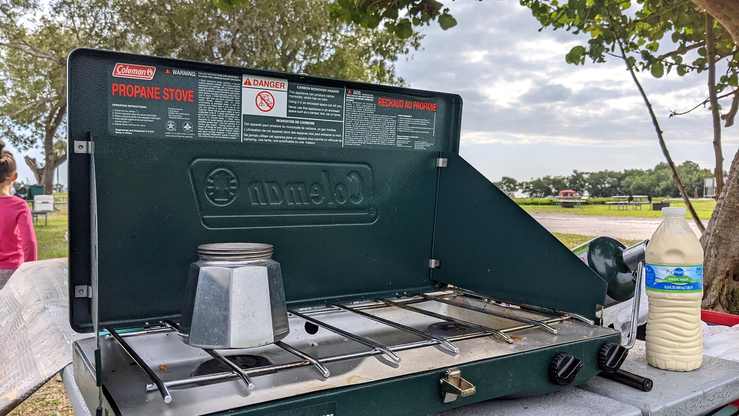 A stovetop moka pot brewing coffee on a green Coleman propane camp stove, with a child in the background at a Florida campsite